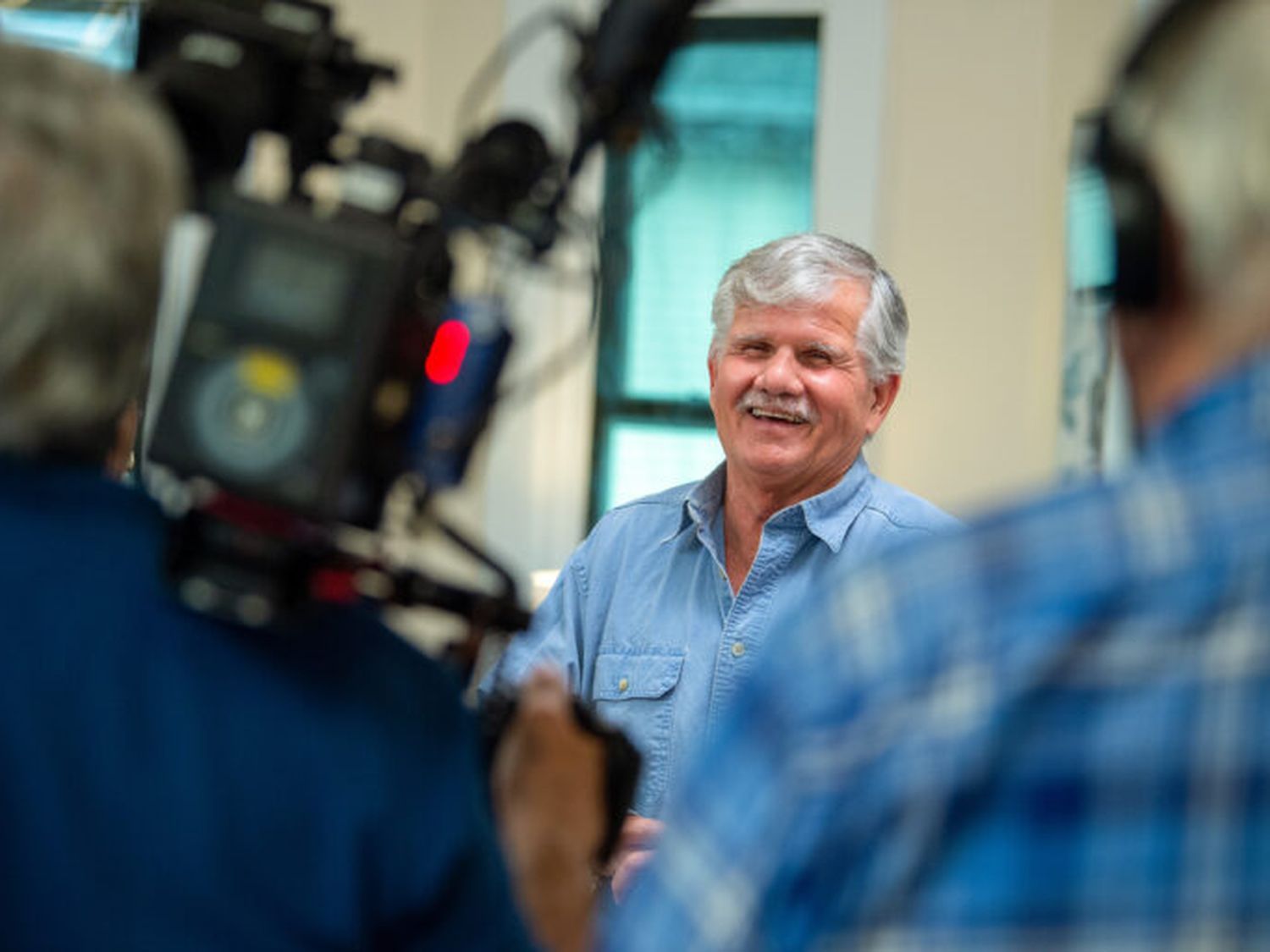 In a cozy room, an older man with gray hair and a mustache smiles while speaking. Dressed in a blue shirt, he shares his insights as the cameraman in blue flannel captures every moment.