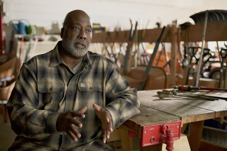 A man with a gray beard, wearing a plaid shirt, sits in a workshop filled with tools and gardening equipment. He gestures with one hand while speaking. A workbench with various tools is in front of him.