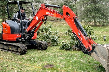 A person operates a small orange excavator with a digging bucket in a grassy area. Nearby, there are some bushes and trees. The ground appears disturbed, as if prepared for construction or landscaping.