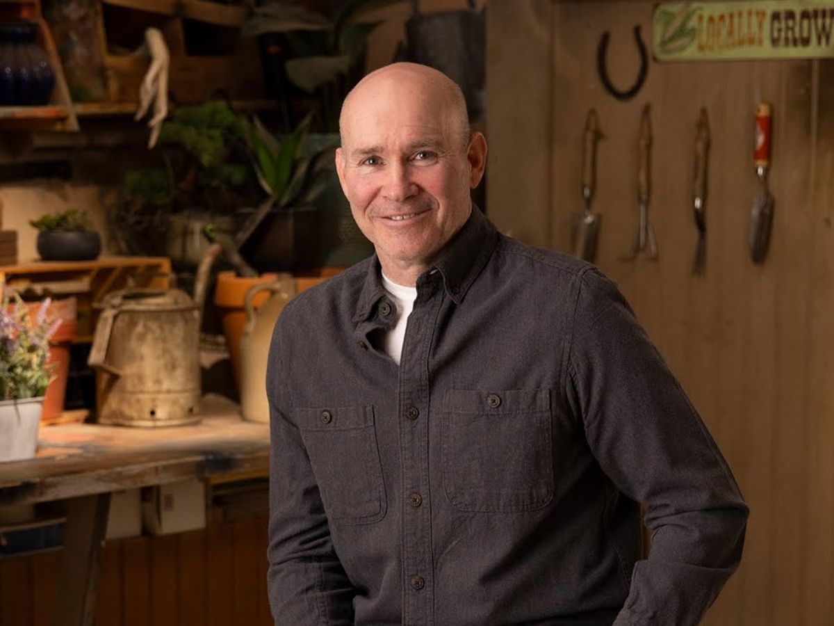 A person in a dark button-up shirt stands smiling in a workshop filled with gardening tools and plants. A wooden shelf with pots, a watering can, and a "Locally Grown" sign is in the background.