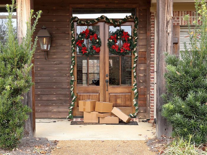 Front porch with wooden double doors adorned with red and green holiday wreaths. Several cardboard packages are stacked by the entrance. Two green bushes flank the doorway, and a brick wall is partially visible on the right.