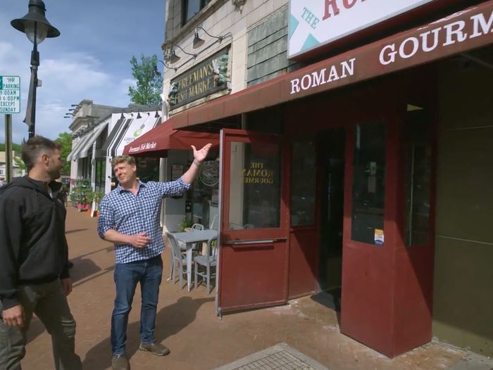 Two people stand outside Roman Gourmet, a red storefront on a street with other shops and outdoor seating. One person gestures upward. A streetlamp and parking sign are visible in the background.