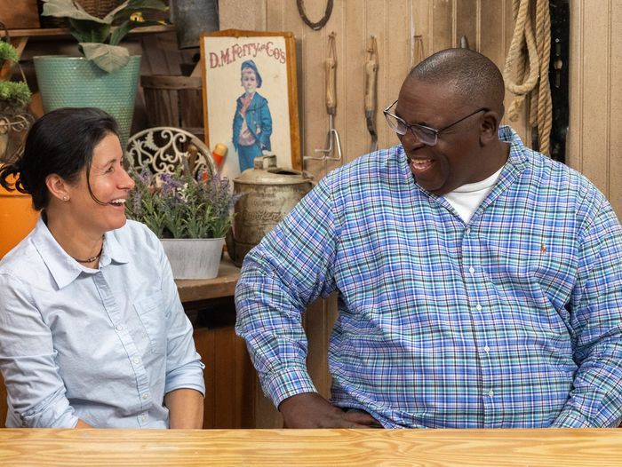 A woman and a man are sitting and laughing together in a cozy, rustic setting. There's a potted plant and a vintage poster in the background. Both are casually dressed and appear to be enjoying a lighthearted conversation.