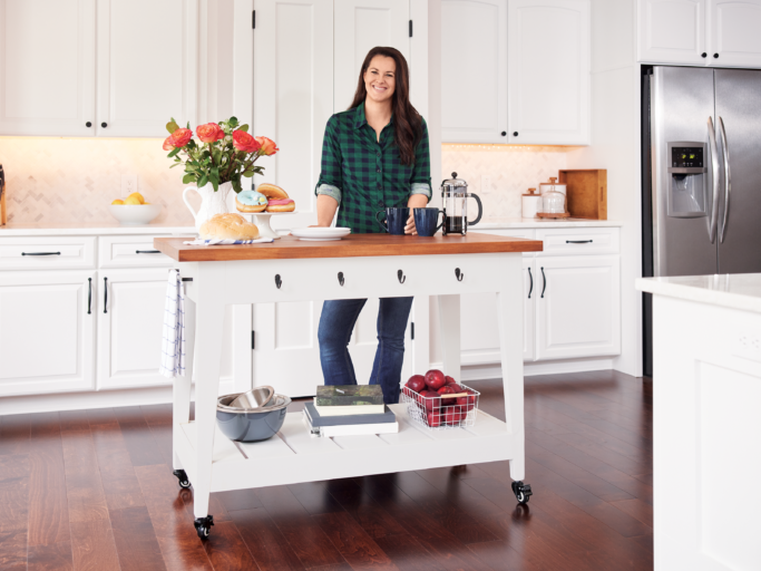 A person stands in a bright kitchen beside a white rolling island with a wooden top. The island holds a bouquet of flowers, loaf of bread, coffee press, and fruit. White cabinets and stainless steel appliances complete the scene.