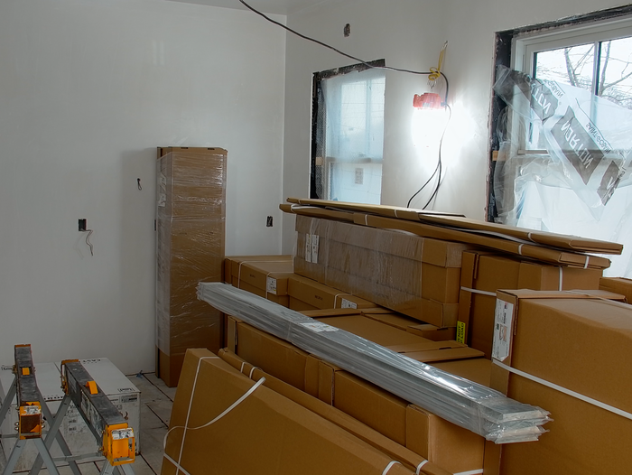 A room under renovation with stacked cardboard boxes, ladders, and plastic-wrapped materials. Walls and windows are partially covered, indicating ongoing construction work. A light illuminates the space, and the floor is unfinished.