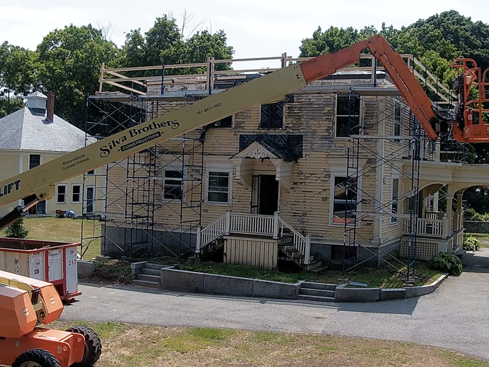 A vintage two-story house under renovation, surrounded by scaffolding. A boom lift with a basket is positioned at the roof level. Trees and another house can be seen in the background.